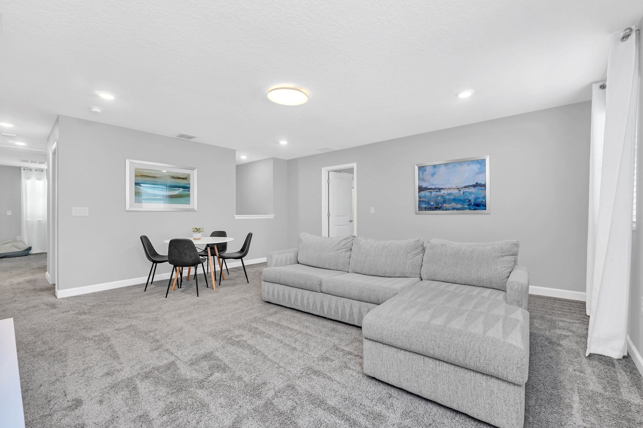 A modern living room with a grey sofa, a dining table with chairs, and two framed paintings on the walls.