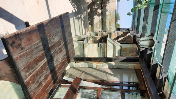 A rustic outdoor seating area with a wooden bench and a window overlooking a stone wall.