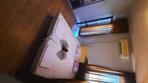 A room with a bed, a window with blinds, a wall-mounted air conditioner, and a door leading to another room.