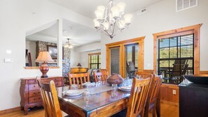 A dining area with a wooden table set for four, surrounded by chairs. A chandelier hangs above the table, and there are windows with blinds on the walls. - Woodland Retreat for 6 | Cozy Fireplace + Pool Access (Branson)