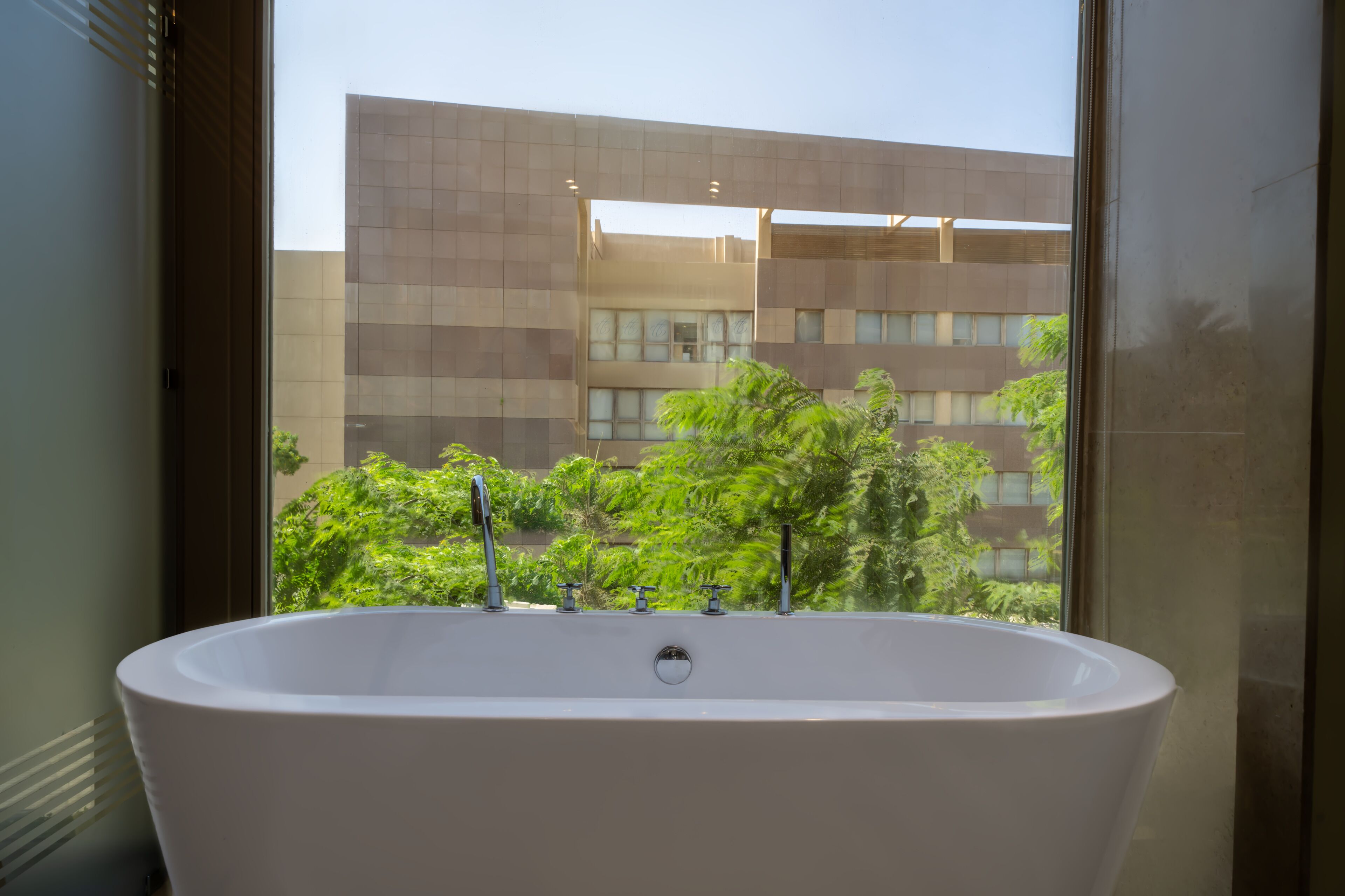 Un baño moderno con una bañera independiente, ventanas amplias y vistas a un edificio y zonas verdes al exterior.