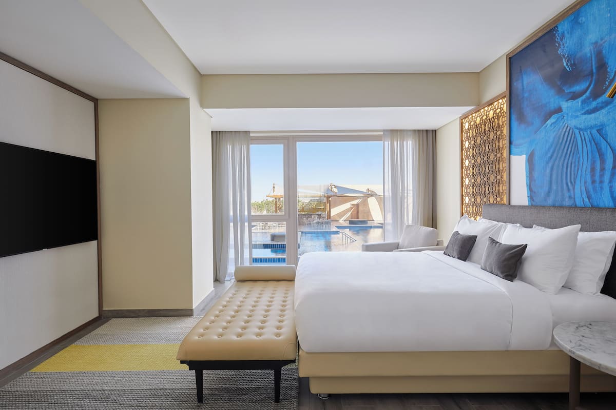 A hotel room with a large bed, a bench, a small table, and a view of a pool and outdoor area.