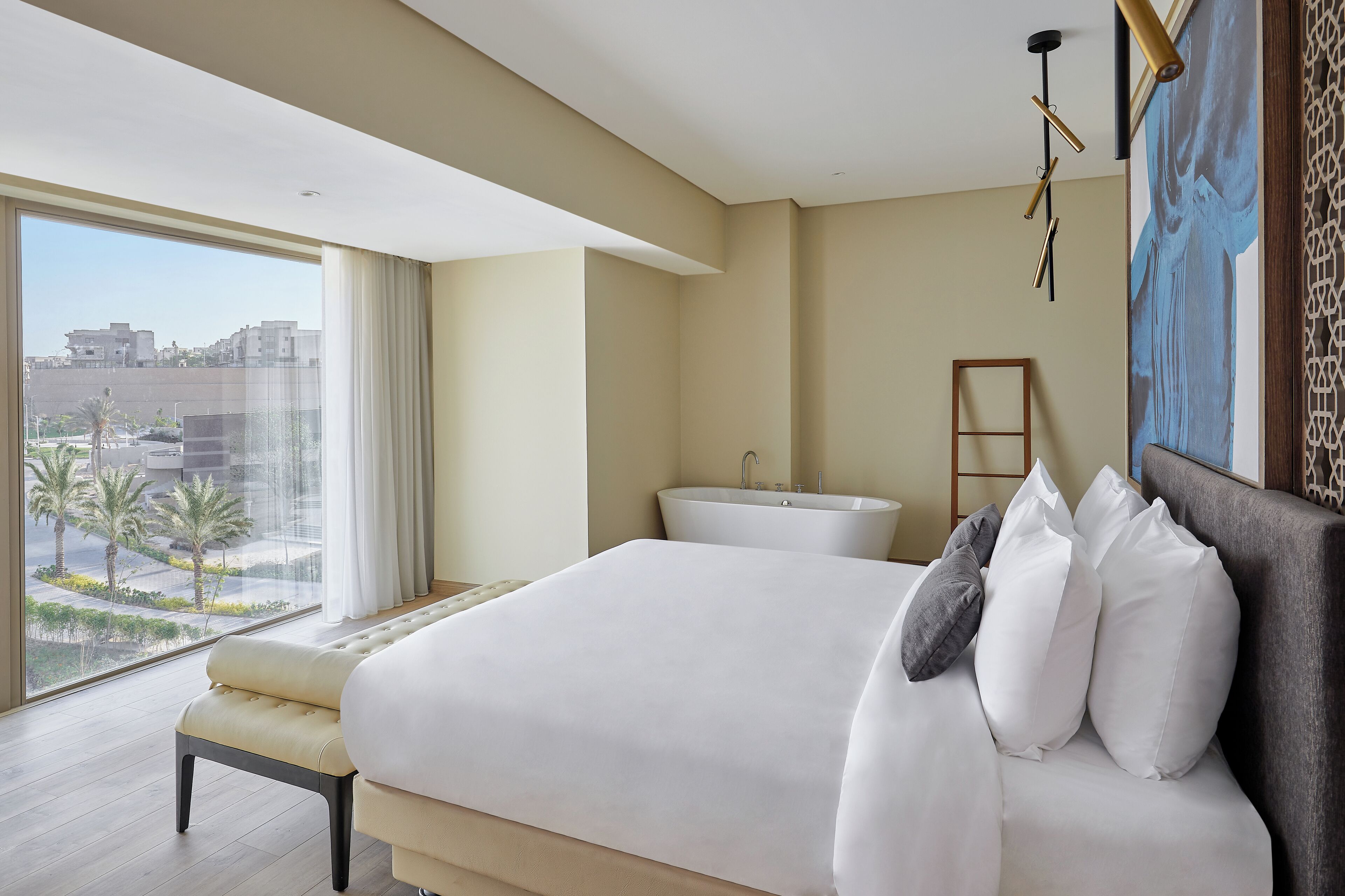 A modern hotel room with a large bed, a view of palm trees and buildings, a bathroom with a bathtub, and a painting on the wall.