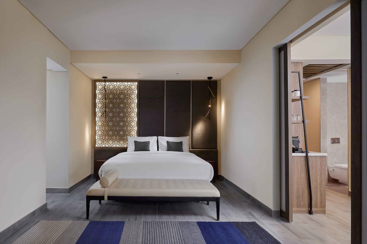 A modern hotel room with a large bed, a bedside table, a bench, a decorative wall panel, and a view of the bathroom.