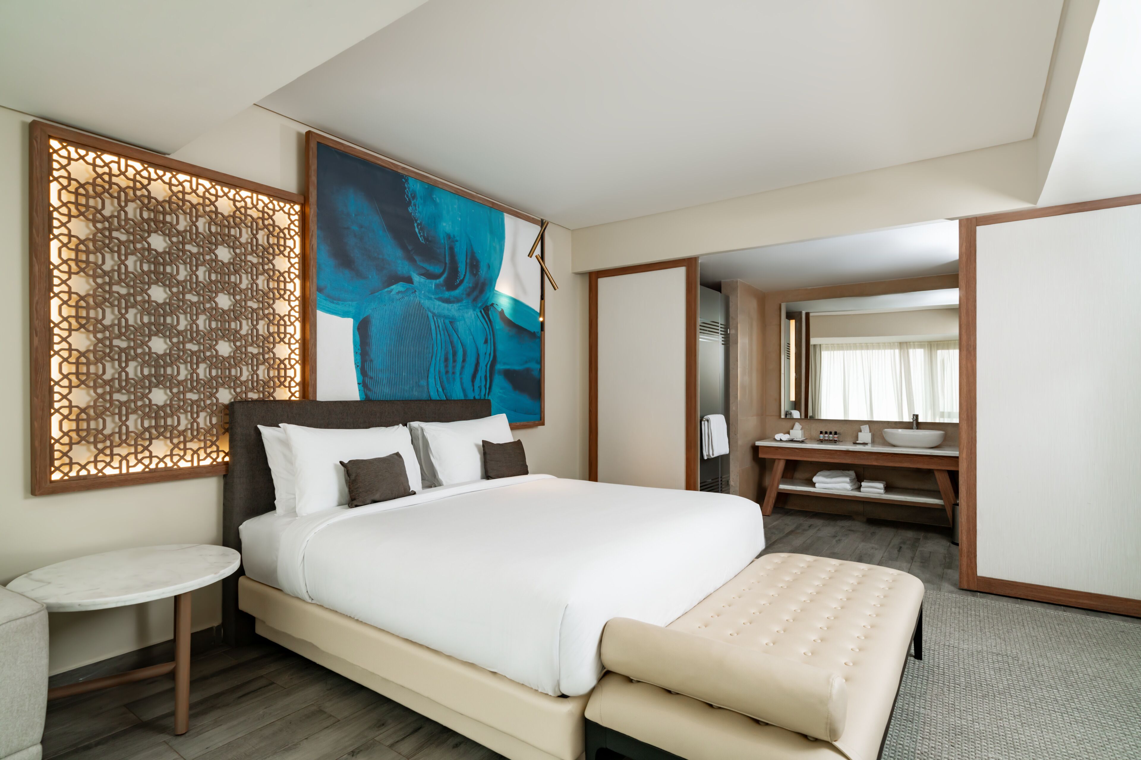 A modern hotel room with a large bed, a round table, a bathroom with a large mirror, and a decorative wall panel.
