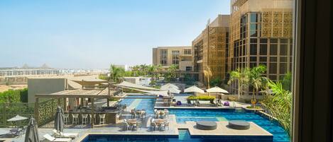 A rooftop pool area with multiple pools, lounge chairs, umbrellas, and a view of the cityscape.