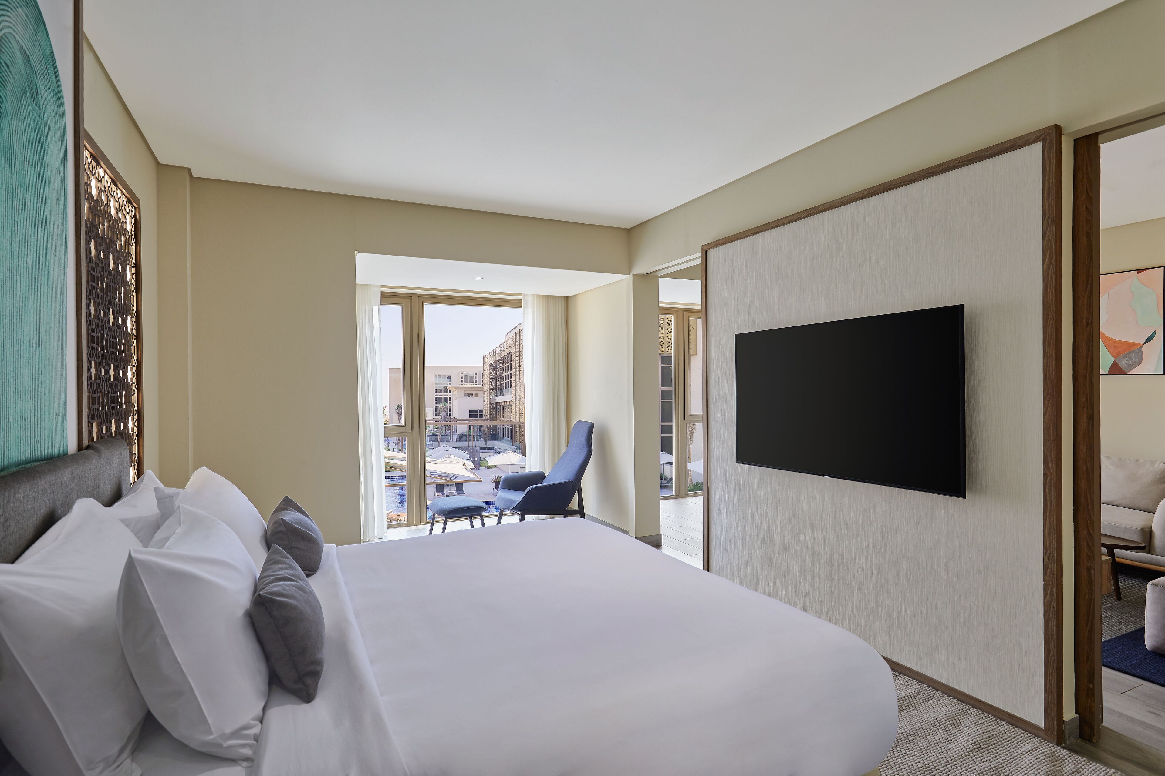 Une chambre d’hôtel dotée d’un grand lit, d’une télévision à écran plat, d’un balcon offrant une vue sur les bâtiments et d’un coin salon avec un fauteuil.