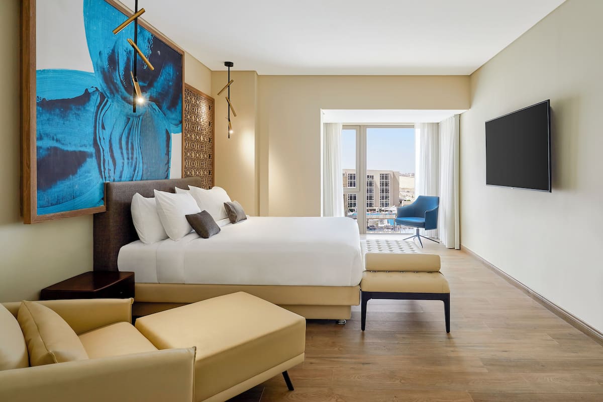 A modern hotel room with a large bed, a sofa, a flat-screen TV, and a balcony with a view of the city.