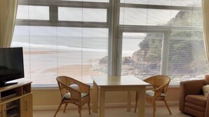 Une pièce dotée d’une grande fenêtre donnant sur une plage, d’un téléviseur posé sur un meuble en bois, de deux fauteuils en osier et d’un canapé beige.