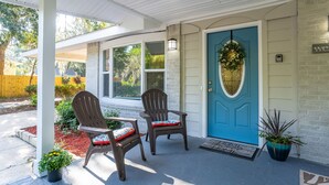 A porch with two Adirondack chairs, a blue door with a wreath, and a potted plant.