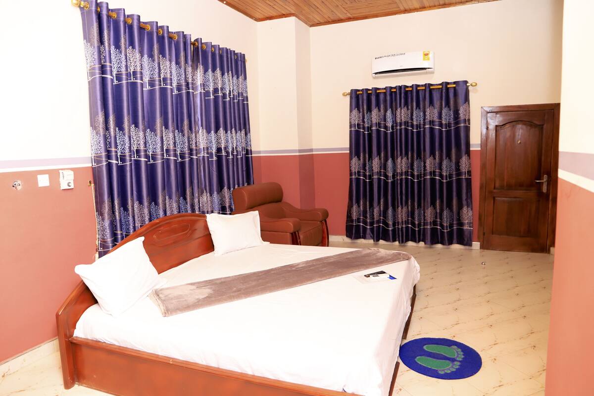 A bedroom with a wooden bed, two blue curtains with tree patterns, a brown chair, a wooden door, and a tiled floor.
