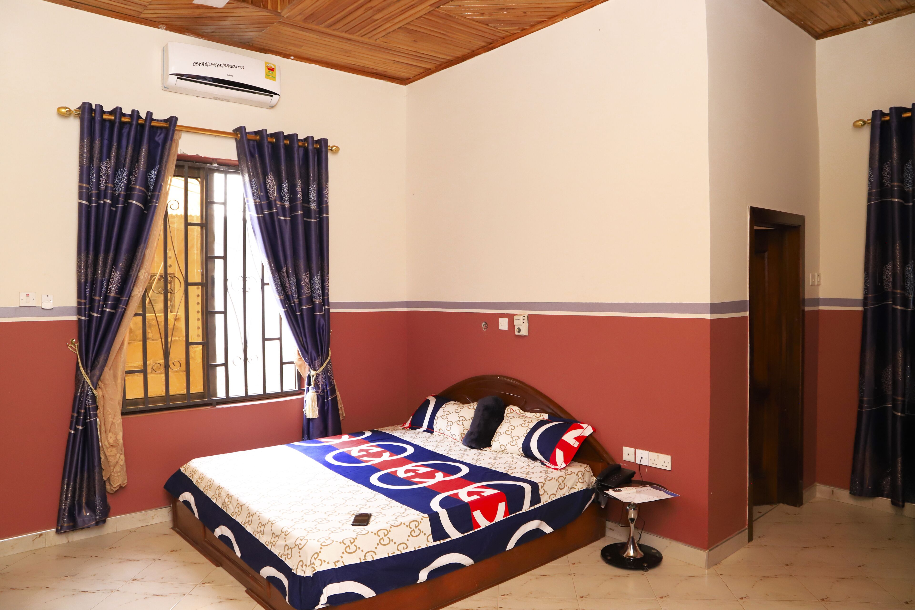 Un dormitorio con cama, cortinas, una ventana con rejas, aire acondicionado y una puerta.