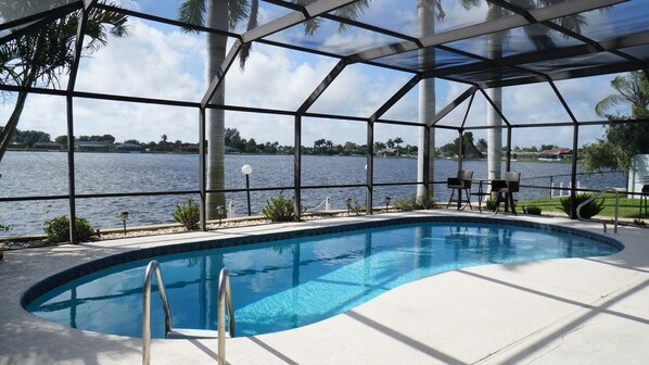 Una piscina con vista a un lago, rodeada por una zona cubierta con sillas y una mesa.