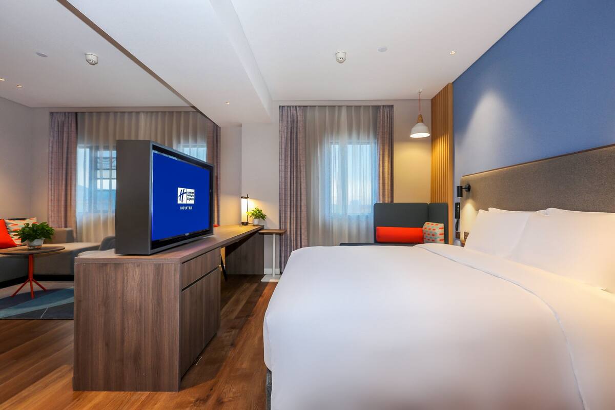 A modern hotel room with a large bed, a desk with a flat-screen TV, and a seating area with a small table and chairs.