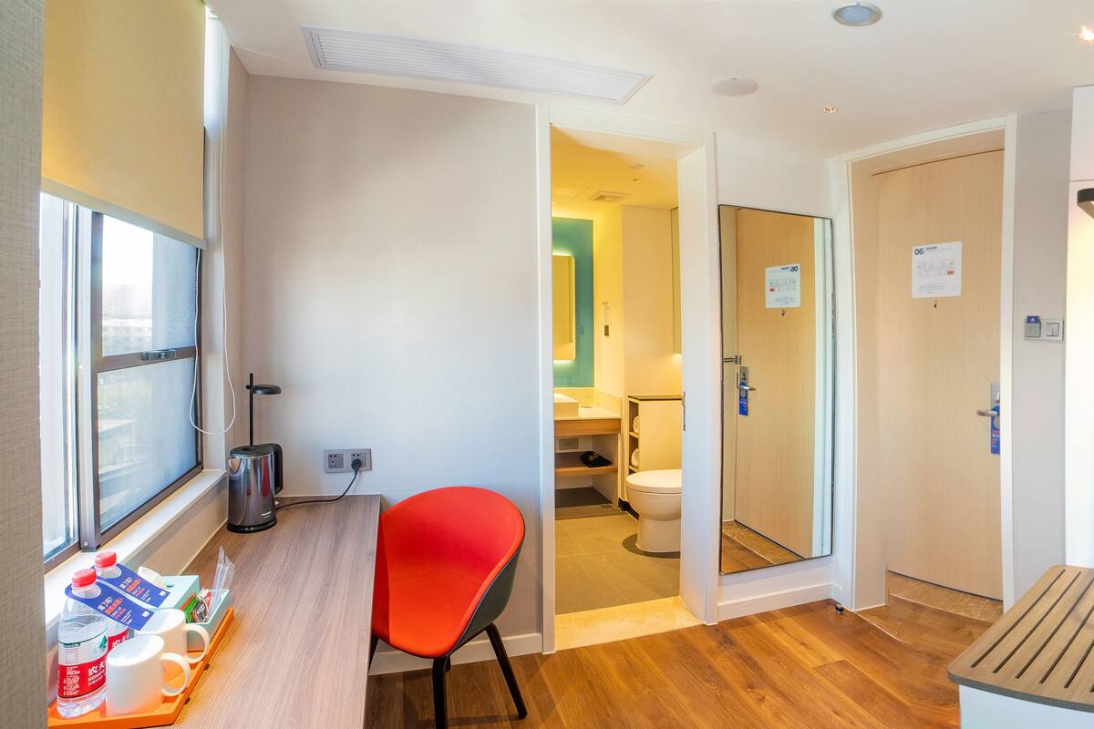 A compact hotel room with a wooden floor, a red chair, a small table with drinks, a window, and a view of the bathroom.