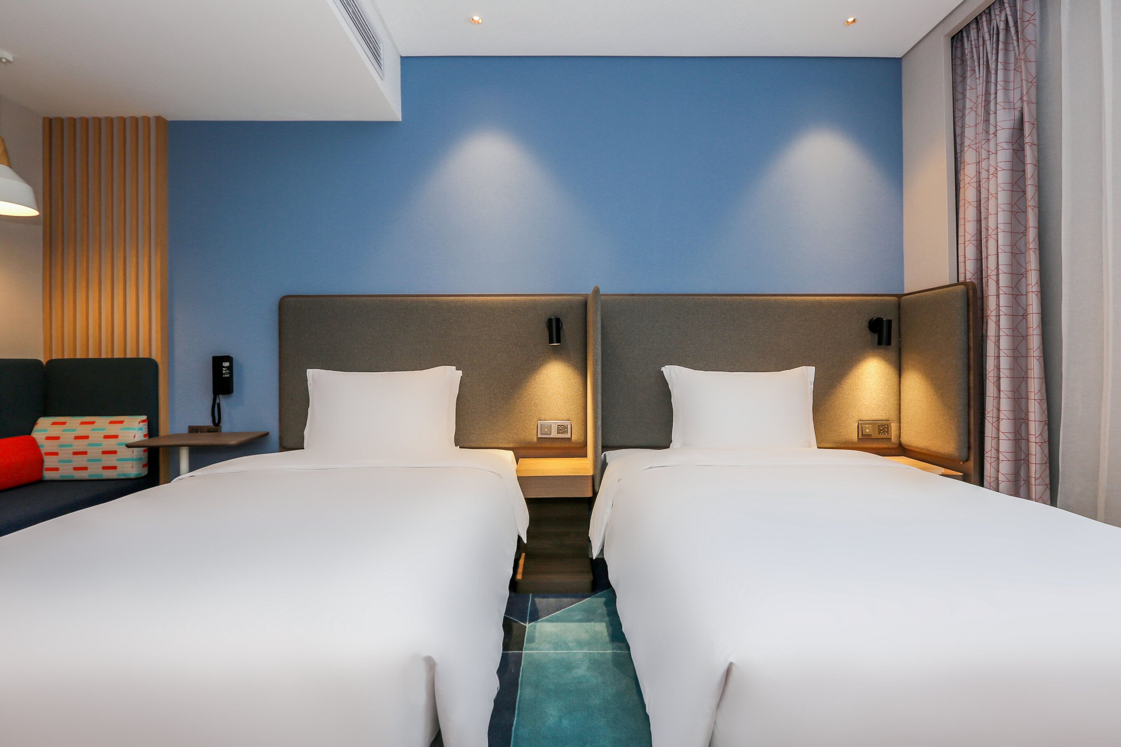 A hotel room with two beds, a small table, a chair, and a window with curtains.