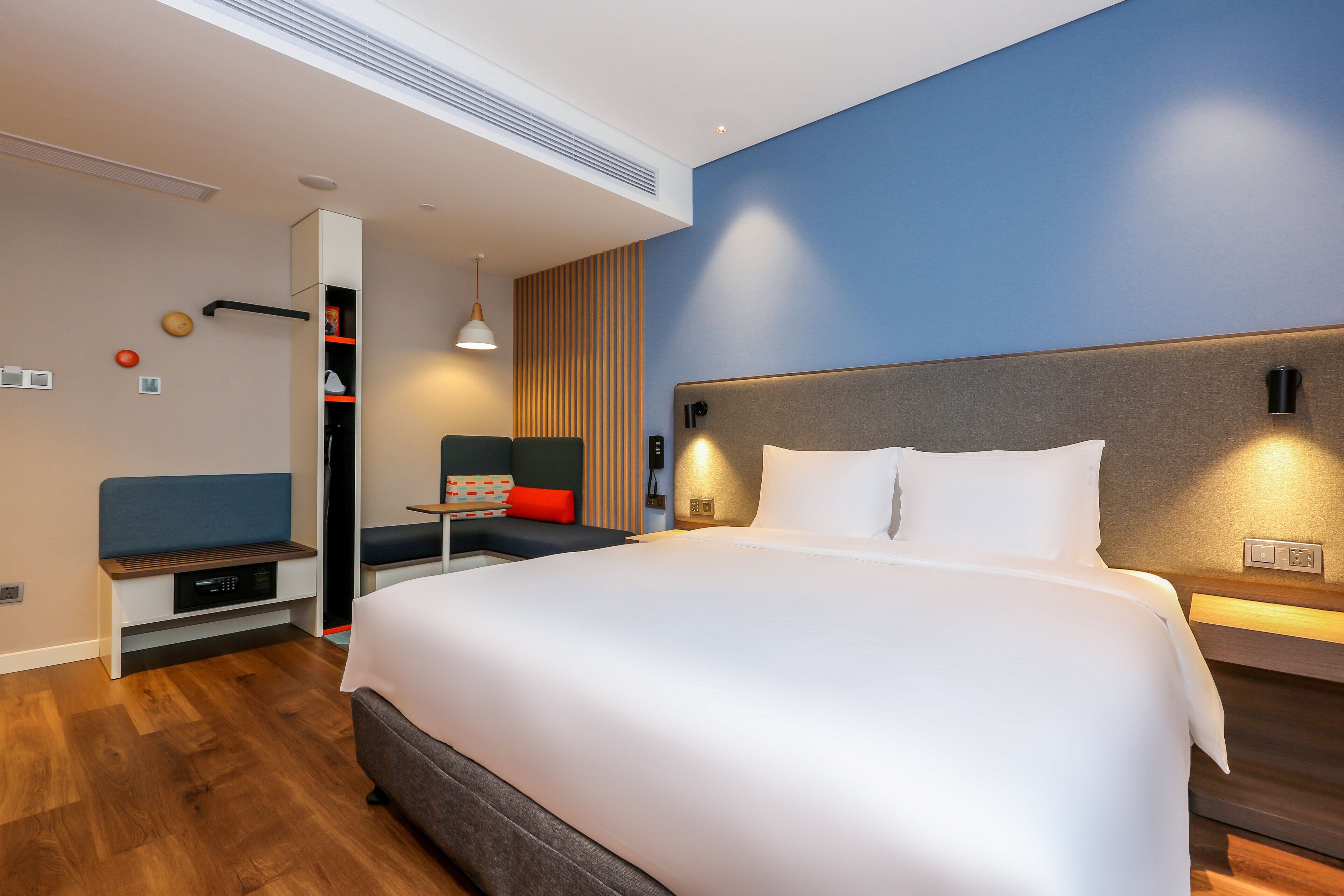 A modern hotel room with a large bed, a seating area with a bench and a chair, and a small table with a lamp.