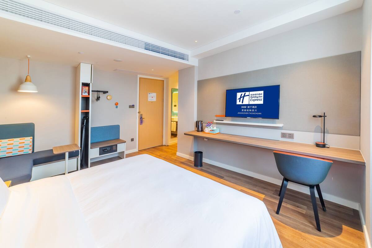 A hotel room with a bed, a desk, a chair, a TV, and a small shelf with toiletries.