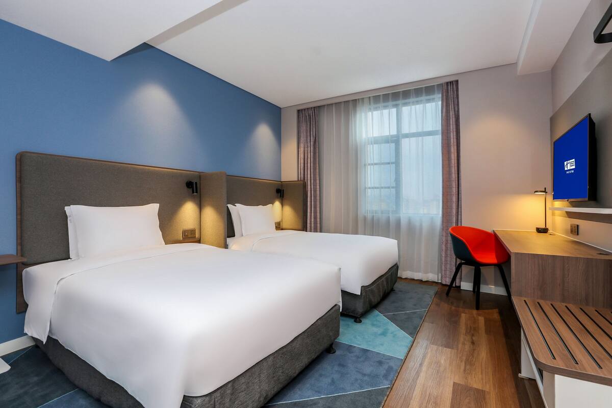 A hotel room with two beds, a desk, a chair, a television, and a window with curtains.