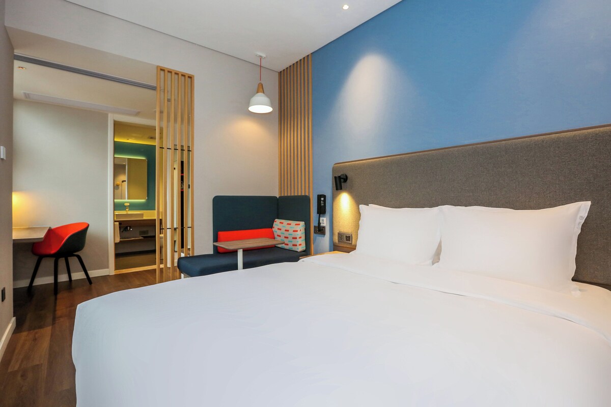 A hotel room with a large bed, a desk with a chair, and a separate seating area with a bench and a chair.