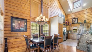 A spacious dining area with a wooden table and chairs, a chandelier, and a kitchen in the background. - Luxurious Walk-in Cabin with Private Screened Deck in the Heart of Branson (Branson)