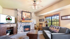 A cozy living room with a stone fireplace, comfortable seating, a television, and a large window offering a view of the outdoors. - Luxurious Walk-in Cabin with Private Screened Deck in the Heart of Branson (Branson)