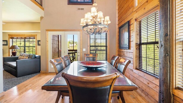 A dining area with a wooden table and chairs, a chandelier, and large windows with blinds. - Luxurious Walk-in Cabin with Private Screened Deck in the Heart of Branson (Branson)