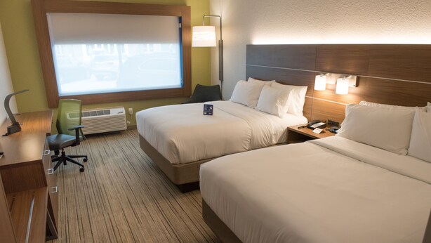 Une chambre d’hôtel avec deux lits, un bureau, une chaise, une lampe et une fenêtre avec des stores.