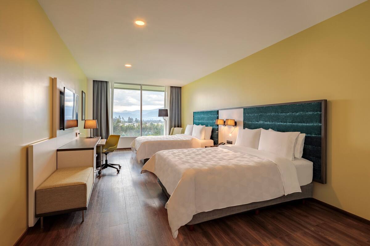 A hotel room with two beds, a desk, a chair, and a view of the mountains.