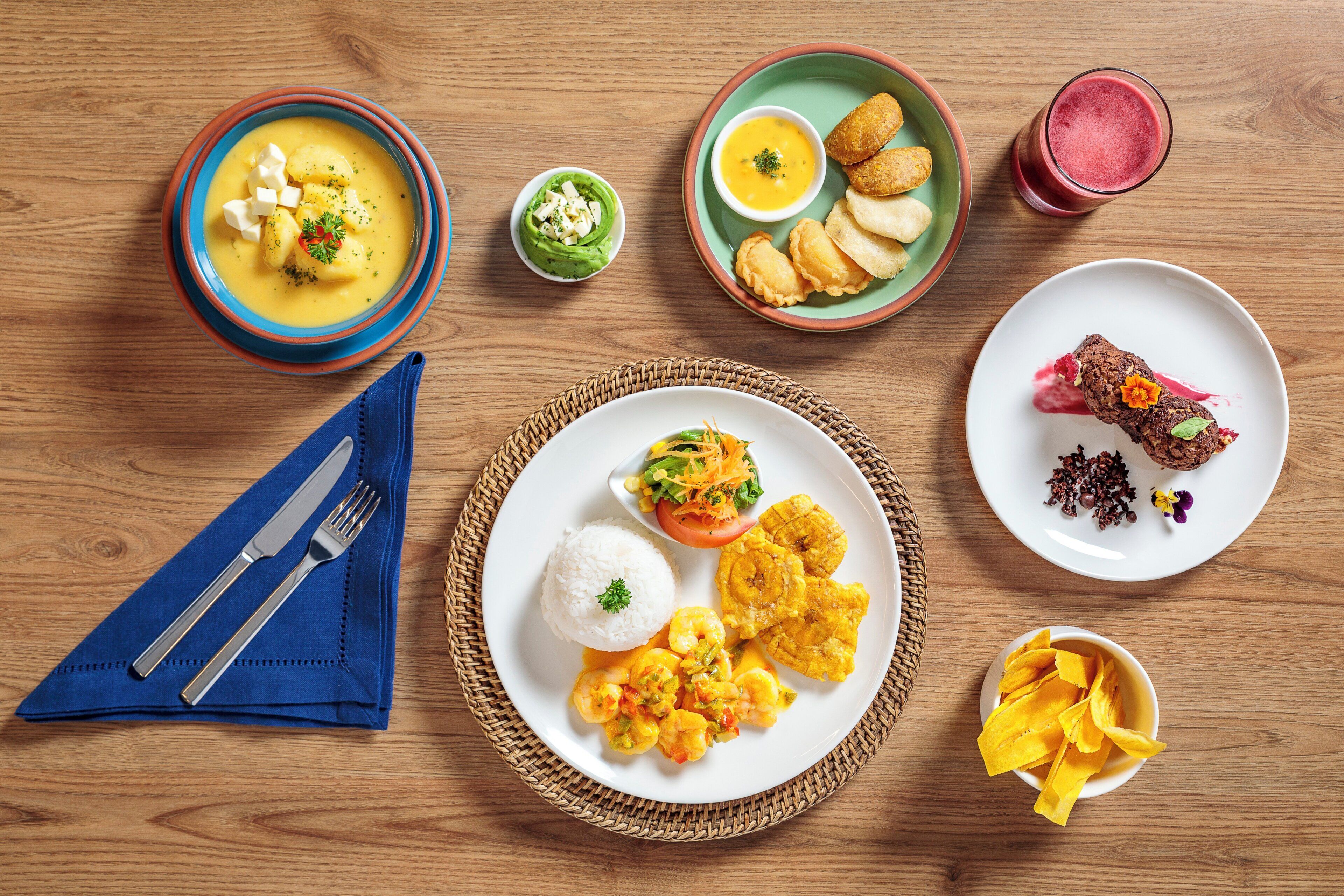 A wooden table with various dishes including soup, rice, shrimp, and plantains.