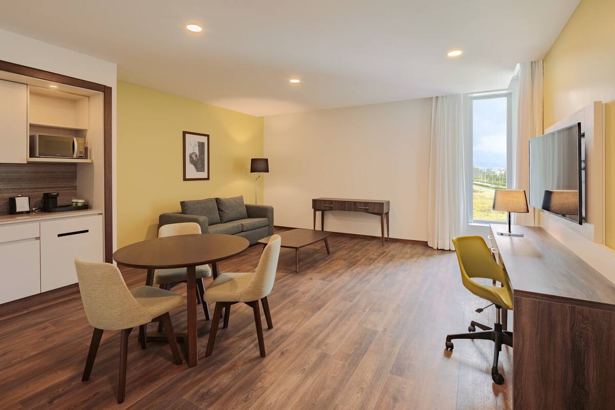 A modern hotel room with a dining area, a sofa, a TV, and a kitchen area with a microwave.