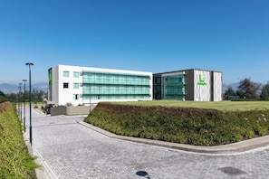 Um edifício moderno com fachada de vidro, telhado plano e área ajardinada com sebes e um caminho pavimentado.