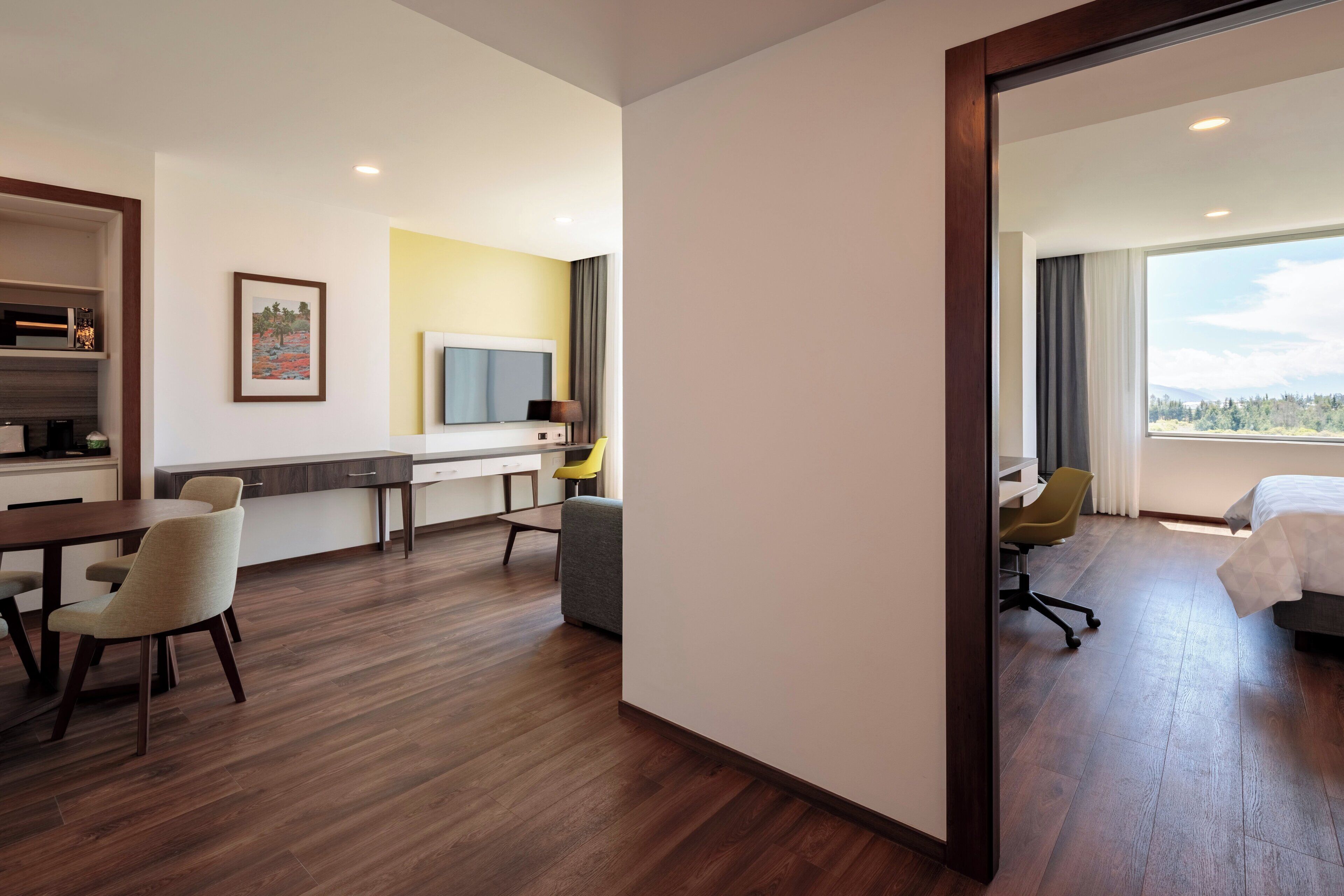 A hotel room with a wooden floor, a desk with a chair, a bed, and a large window with curtains.