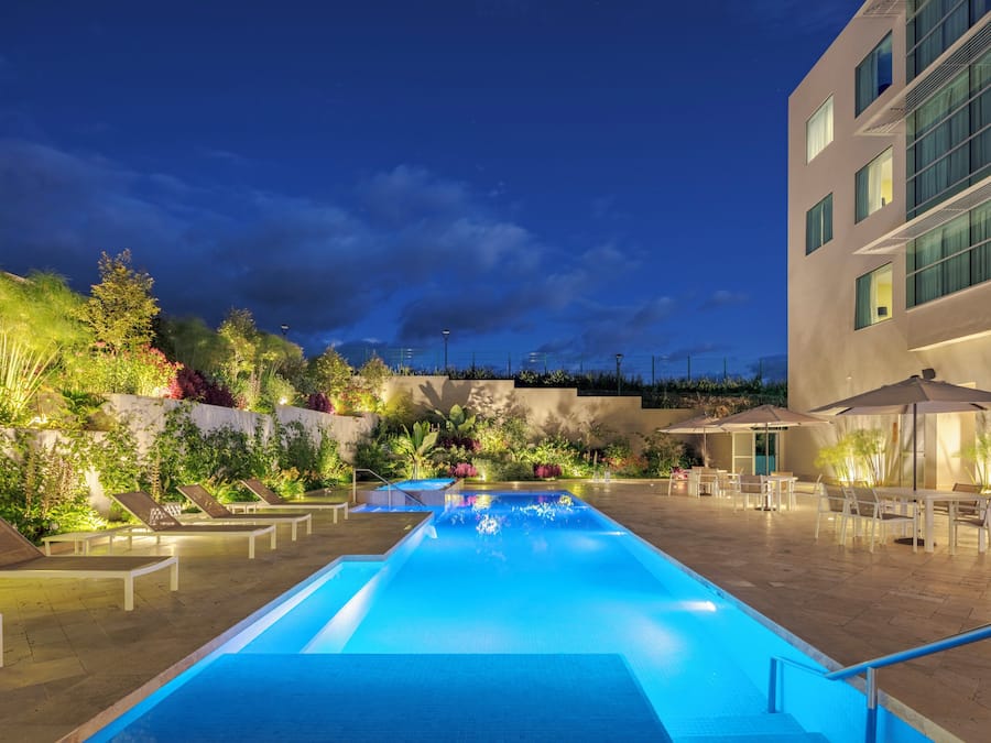 Una piscina con sillas de descanso y sombrillas, rodeada de vegetación y un edificio.