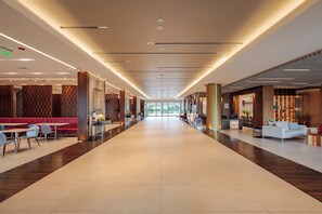 Corredor de hotel espaçoso com design moderno, apresentando um caminho central, áreas de estar em ambos os lados e uma receção no final.