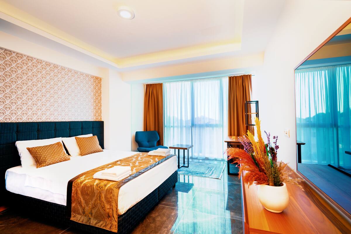 A hotel room with a large bed, a small table, a chair, and a view of the city through large windows.