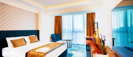 A hotel room with a large bed, a small table, a chair, and a view of the city through large windows.