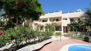 Un hotel de varios pisos con zona de piscina, rodeado de vegetación exuberante y palmeras.