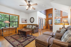 A spacious living room with a ceiling fan, wooden floors, a large window, and a cozy seating area with a sofa and ottoman. - "Woodsy" Walk-in Cabin near Everything Branson has to Offer! (Branson)
