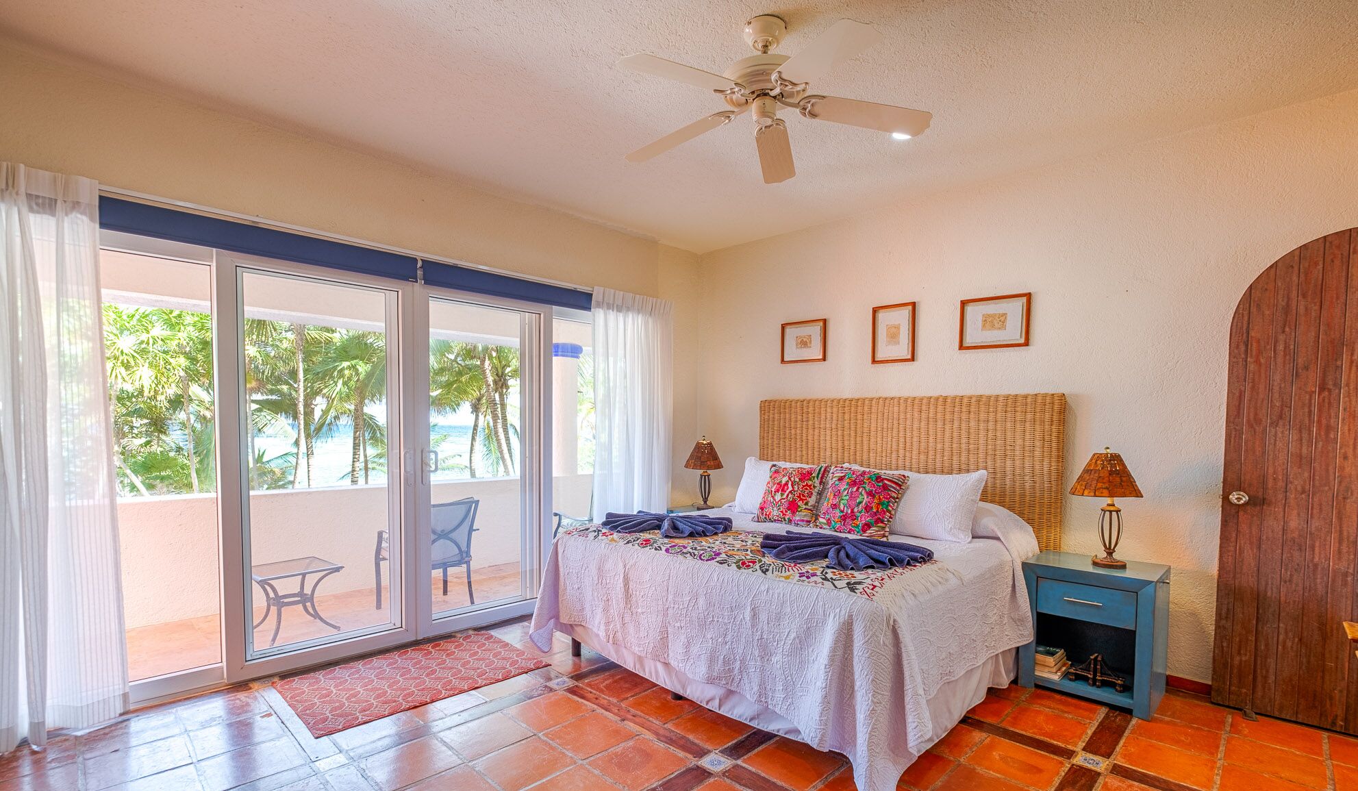 A bedroom with a bed, a ceiling fan, a nightstand, a lamp, a wooden door, and a sliding glass door leading to a balcony with a chair and a table.