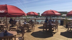 An outdoor dining area with wooden tables and chairs, red umbrellas, and a pool in the background.