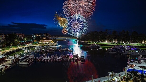 A marina with boats docked, a waterfront promenade, and a large fireworks display in the background.
