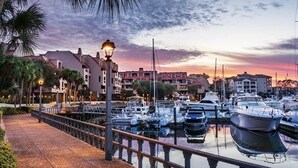 A marina with boats docked, a waterfront promenade, and buildings in the background.