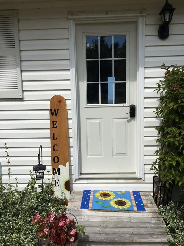 Una casa blanca con un letrero que dice 'WELCOME', una puerta y un felpudo azul con girasoles.