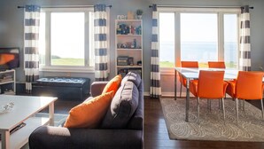 A living room with a sofa, ottoman, and a dining area with a table and chairs. There are windows with curtains, a bookshelf, and a view of the sea. - Fundy Sunset Cottage, Oceanfront Tranquility!! (granville ferry)