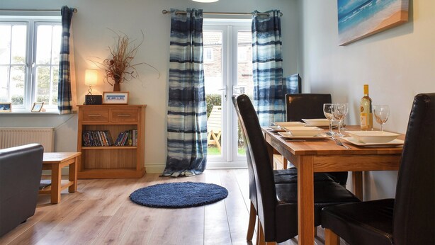 A dining area with a wooden table, chairs, a blue rug, a bookshelf, and a window with curtains. - 2 bedroom accommodation in Filey (Filey)