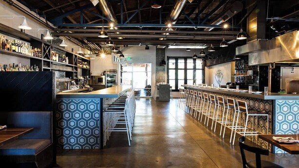 Una zona de bar con una barra larga, diversas opciones de asiento y una cocina a la vista al fondo.
