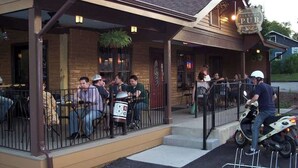 Un área de estar al aire libre de un pub con mesas y sillas, un porche cubierto y una persona en un scooter.