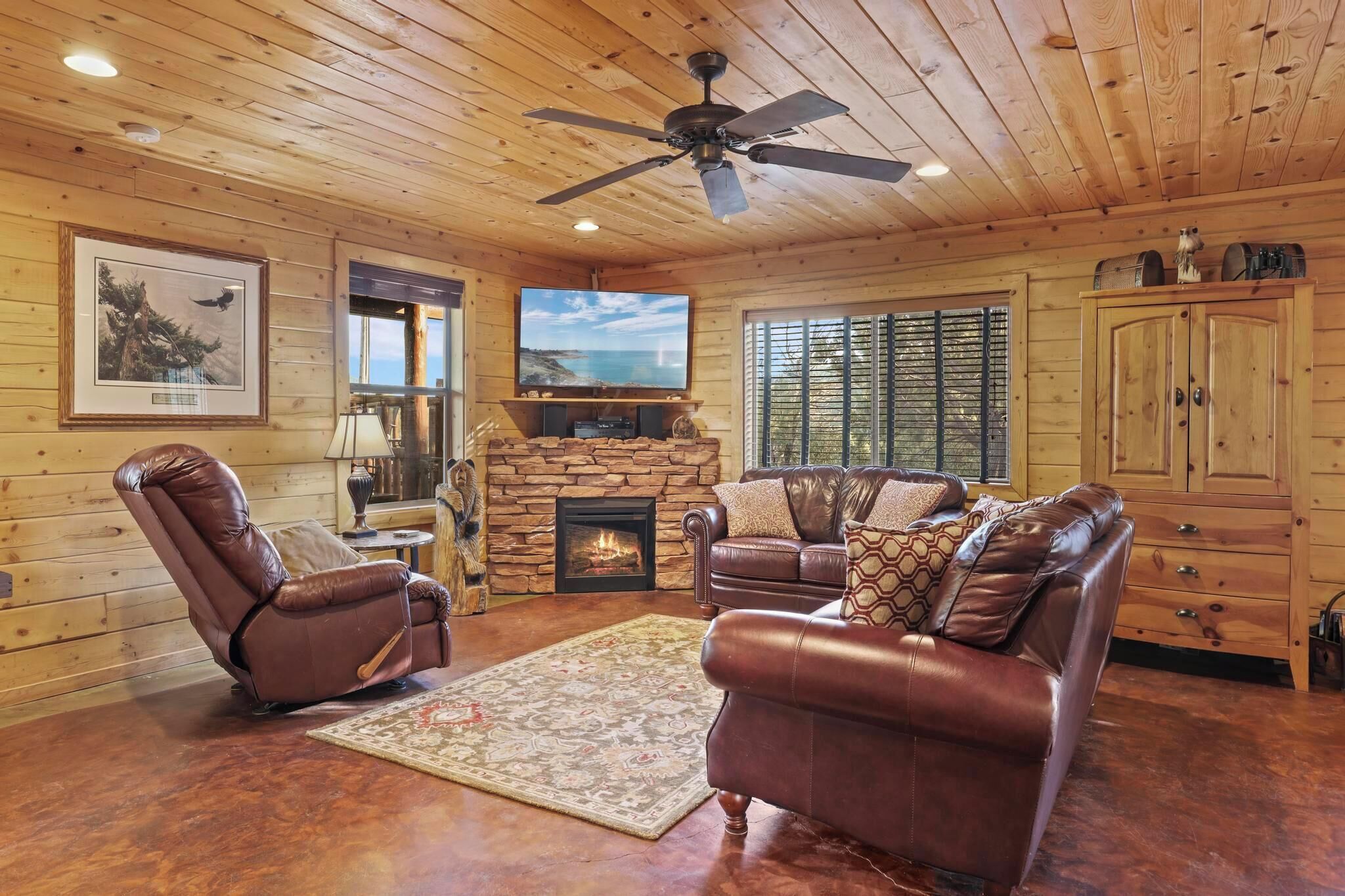 A cozy living room with a fireplace, leather sofas, a ceiling fan, and wooden walls.