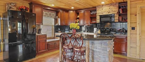 A kitchen with wooden cabinets, a black refrigerator, a stone island, and two bar stools.